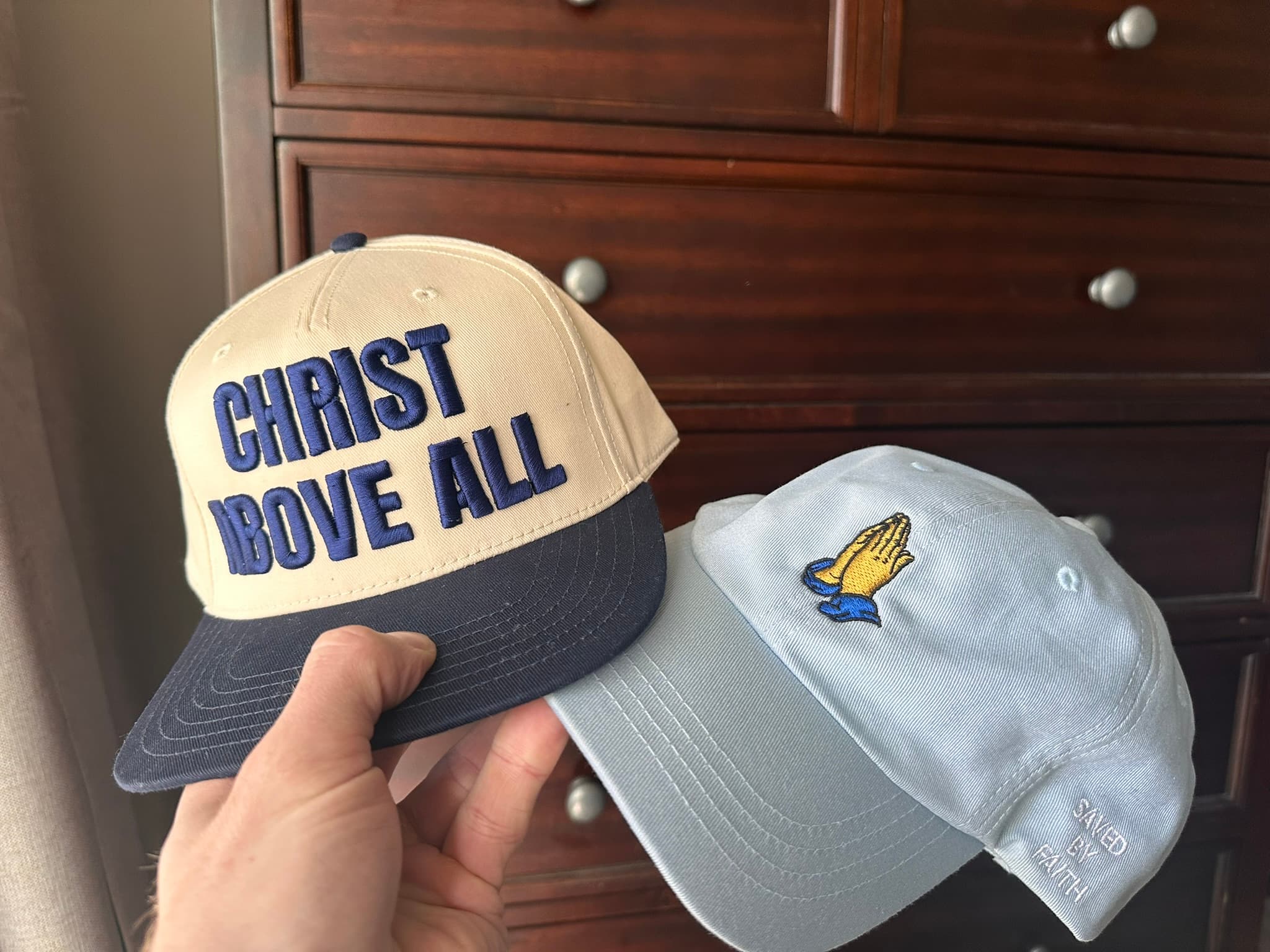 Saved by Faith hats in natural and light blue held in front of a wooden cabinet